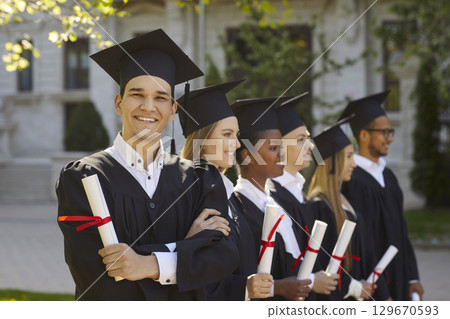Happy graduate man in graduation gown and cap with diploma posing for photo standing near students. 129670593