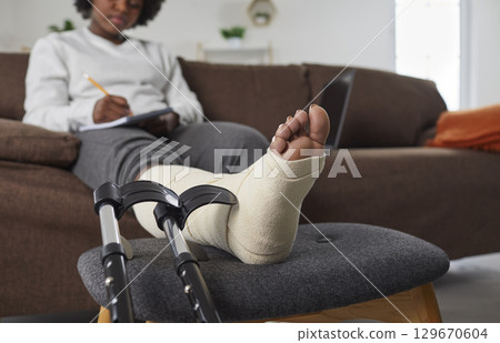African American woman with broken leg sitting on sofa, with her foot resting on stool 129670604