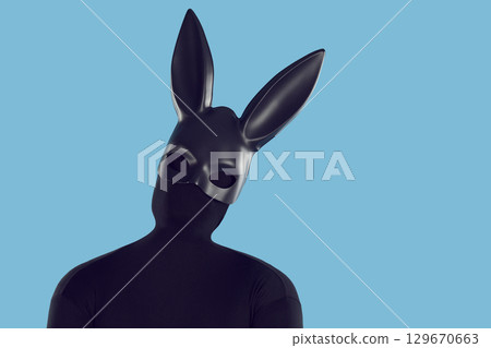 Man disguised in black bodysuit and long eared bunny mask tilts head on blue background 129670663