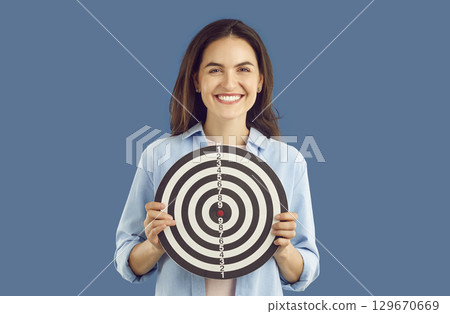 Positive smiling brunette woman in casual shirt holding darts shooting target on blue background. 129670669