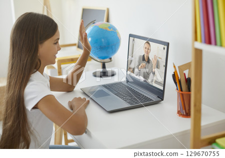 Happy school girl learning online, having remote class and waving hand at laptop screen 129670755