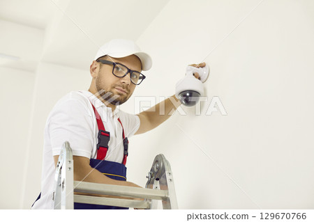 Portrait of maintenance worker adjusting wall-mounted dome surveillance camera inside house. 129670766