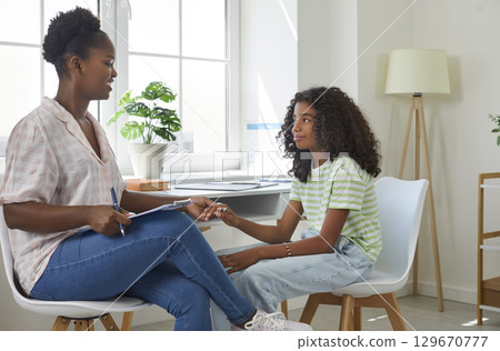 Smiling afro american woman psychologist holding hand teen girl sitting near on therapy session. 129670777