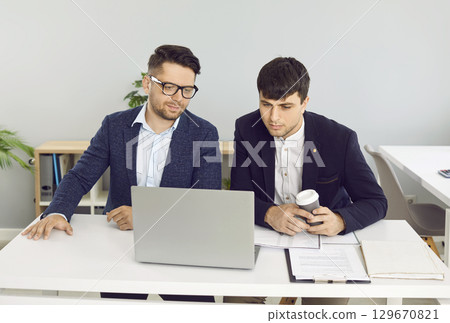 Two young businessmen sitting at an office desk and looking at their laptop computer 129670821
