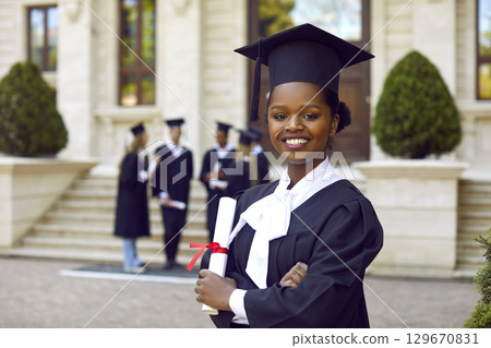 Happy smiling afroamerican woman in graduation gown and cap graduates universuty looking at camera. Happy smiling afroamerican woman in graduation gown and cap graduates universuty looking at camera. 129670831