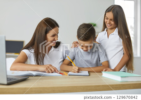 Child boy is learning to write in notepad sitting at desk with his mom and sister looking at him. Child boy is learning to write in notepad sitting at desk with his mom and sister looking at him. 129670842
