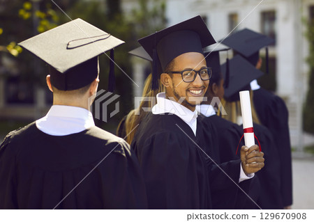 Half turned indian graduate man in graduation clothes looking at camera and smiling on ceremony. 129670908