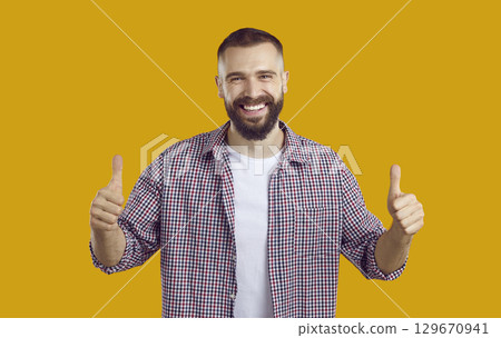 Young brunet bearded man in plaid shirt showing thumbs up on yellow background looking at camera. Young brunet bearded man in plaid shirt showing thumbs up on yellow background looking at camera. 129670941