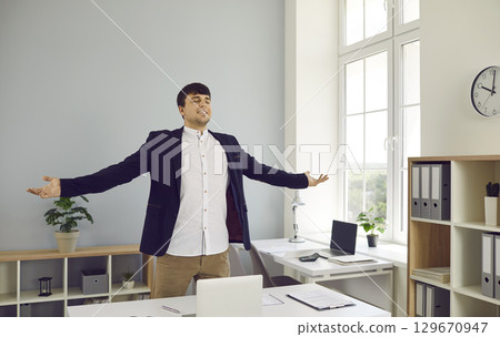 Young man office worker stands next to work desk with business documents, spreading arms to sides Young man office worker stands next to work desk with business documents, spreading arms to sides 129670947