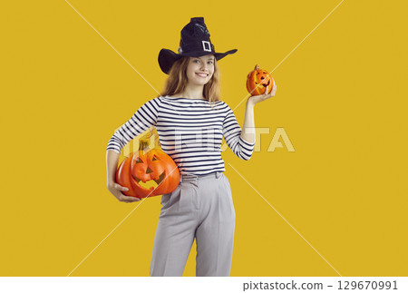 Young attractive student-age woman holds two Halloween pumpkins stands on orange studio background 129670991