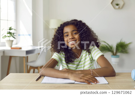 Smiling african american teen girl is studying online looking at camera sitting at desk at home. 129671062