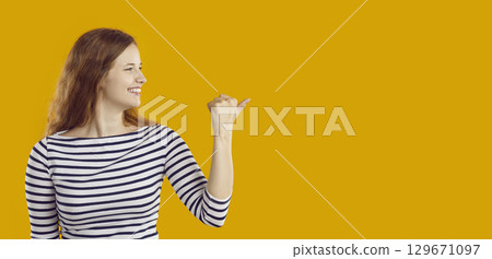Cheerful cute young woman pointing with thumb at place for announcements or advertisements. Cheerful cute young woman pointing with thumb at place for announcements or advertisements. 129671097