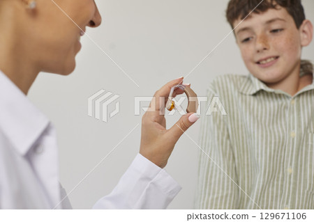 Modern effective hearing aid in hands of female ENT doctor showing it to smiling preteen boy. 129671106