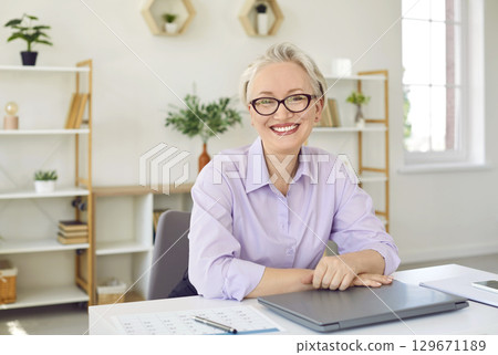 Gray haired elderly Caucasian woman smiling looking at camera sits at home office desk with laptop 129671189