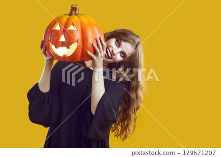Woman with scary make-up dressed in black gown is holding Halloween pumkin on yellow background. 129671207