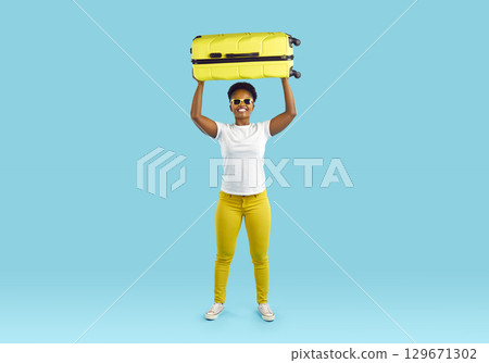 Full length portrait of woman with yellow suitcase over her head smiling on turquoise background. 129671302