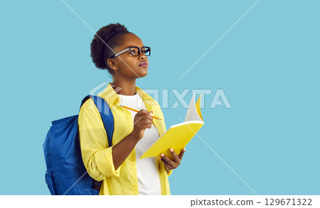 Student girl standing on blue background, holding pencil and notebook, writing essay and thinking 129671322