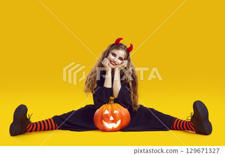Happy woman in Halloween costume sitting on yellow studio background with Jack o lantern Happy woman in Halloween costume sitting on yellow studio background with Jack o lantern 129671327