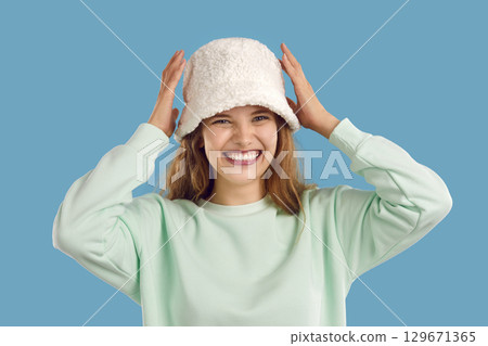 Portrait of laughing girl in white warm fluffy hat and mint sweatshirt on light blue background. Portrait of laughing girl in white warm fluffy hat and mint sweatshirt on light blue background. 129671365