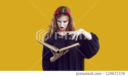 Young girl in carnival costume of devil holding book in hands and conjuring in orange background. Young girl in carnival costume of devil holding book in hands and conjuring in orange background. 129671376