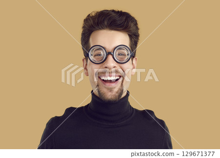 Funny young man in glasses with magnified lenses laughs isolated on beige background. Funny young man in glasses with magnified lenses laughs isolated on beige background. 129671377