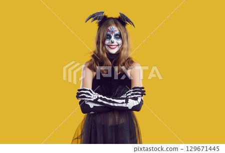 Smiling girl in creative skeleton costume and bat wings on her head with skull makeup, crossed her arms over her chest. 129671445