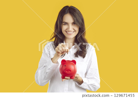 Portrait of satisfied happy woman who throws coin into piggy bank to save money. 129671455