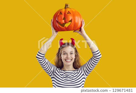 Happy young woman with devil horns holding up orange Halloween pumpkin and smiling Happy young woman with devil horns holding up orange Halloween pumpkin and smiling 129671481