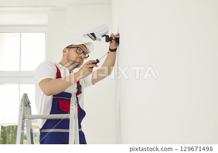 A professional worker standing on a stepladder installs a video surveillance camera on the wall. 129671498
