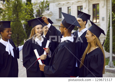 Young guy adjusts mortar boards cap his smiling blonde classmate. 129671517