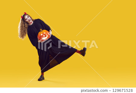 Evil girl in devilish costume dances with pumpkin isolated on yellow background. 129671528