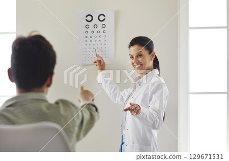 Smiling cheerful female ophthalmologist checking vision of little boy with eye test board. Smiling cheerful female ophthalmologist checking vision of little boy with eye test board. 129671531