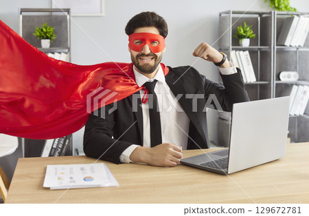 Businessman in suit, red superhero mask and cape, showing his arm muscles seated at desk with laptop 129672781