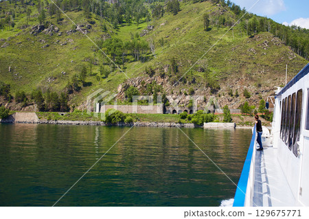 Summer cruise. A boy on the deck of a ship admires nature. The shore of Lake Baikal, mountains, forest.  129675771
