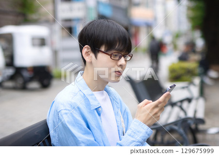 一名年輕人坐在城市的長椅上，看著他的智慧型手機 129675999