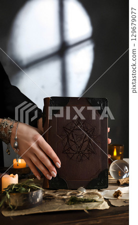 Female hands take old grimoire from a table with magic tools, vertical photo, copy space. 129679077