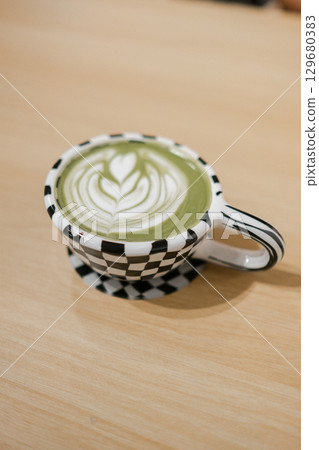 A beautifully crafted matcha latte with heart-shaped latte art in a stylish checkered cup on a wooden table. 129680383