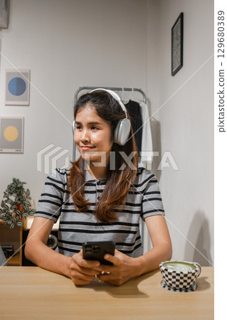 A young woman wearing headphones, holding a smartphone, sitting at a table in a modern minimalist room with framed art and plants. 129680389