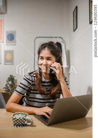 Smiling woman using a laptop and phone in a cozy home office environment, showcasing multitasking and communication. Smiling woman using a laptop and phone in a cozy home office environment, showcasing multitasking and communication. 129680395