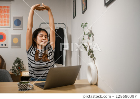 A woman stretches at her home office desk, surrounded by art and plants, promoting relaxation and productivity. A woman stretches at her home office desk, surrounded by art and plants, promoting relaxation and productivity. 129680397