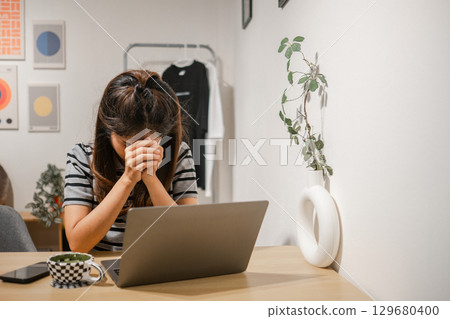 A woman sits at a desk with a laptop, appearing stressed, in a modern home office environment. 129680400