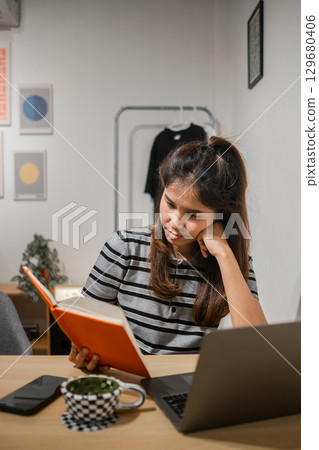 A young woman reads a book at her home office desk, surrounded by a laptop and smartphone, creating a cozy and modern workspace atmosphere. A young woman reads a book at her home office desk, surrounded by a laptop and smartphone, creating a cozy and modern workspace atmosphere. 129680406