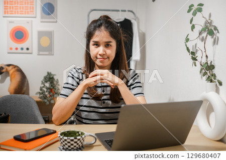 Young woman in a home office setting with a laptop, smartphone, and modern decor, exuding confidence and focus. 129680407