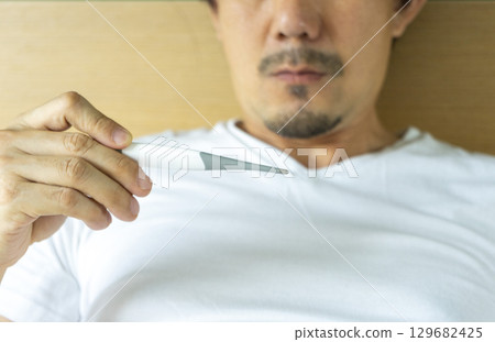 Sick man with fever laying in bed taking temperature with thermometer Sick man with fever laying in bed taking temperature with thermometer 129682425
