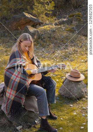 Blonde woman playing string guitar outdoors in autumn forest. Concept of sound therapy, mental health and wellness rituals. Calmness tranquility audio-sensory practices. Aura farming energy 129682503