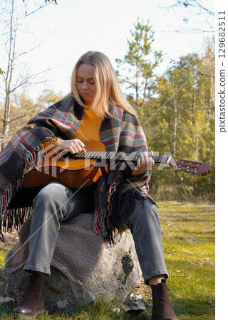 Beautiful blonde girl playing acoustic guitar and singing relaxing enjoying outdoors on sunny autumn day. Hippie woman in boho style learning and practicing playing musical instrument. Fall leaves Beautiful blonde girl playing acoustic guitar and singing relaxing enjoying outdoors on sunny autumn day. Hippie woman in boho style learning and practicing playing musical instrument. Fall leaves 129682511