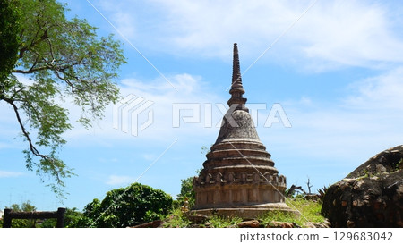 An ancient, weathered stupa (chedi) standing amongst rocks and green trees against a bright blue sky, showcasing historical Buddhist architecture in Thailand An ancient, weathered stupa (chedi) standing amongst rocks and green trees against a bright blue sky, showcasing historical Buddhist architecture in Thailand 129683042
