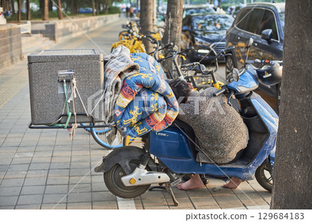 A person resting on a blue scooter parked on a sidewalk, with a blanket and a box attached to the scooter, in Beijing, China 129684183