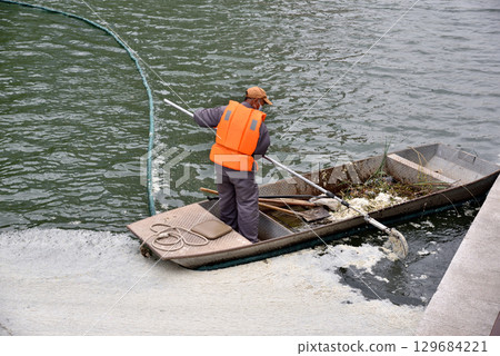 Worker in orange vest in a small boat cleaning debris from water of Liangma river in central Beijing, China 129684221