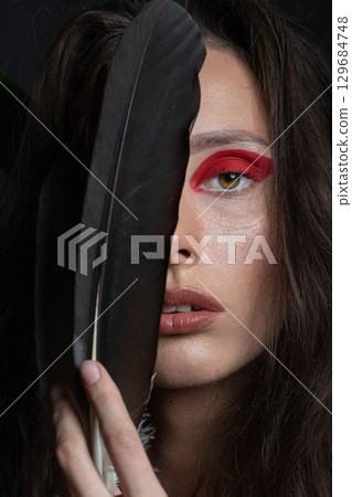 Studio portrait of woman with red eyes shadows covering one eye with big bird feather 129684748